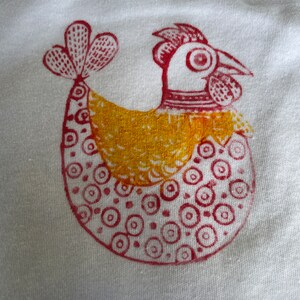 Op de afbeelding: Een handgeprint ontwerp van een kip op een witte stoffen achtergrond. De kip is rood en geel, met een rode kam en rode keellap, een geel lichaam en een rode staart.