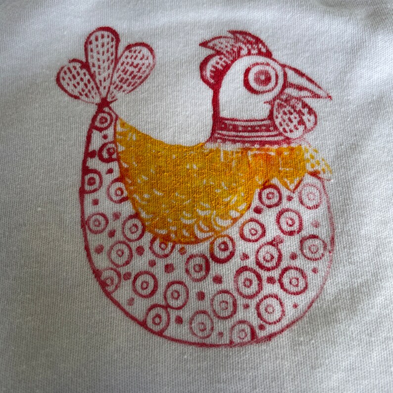 Op de afbeelding: Een handgeprint ontwerp van een kip op een witte stoffen achtergrond. De kip is rood en geel, met een rode kam en rode keellap, een geel lichaam en een rode staart.