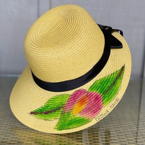 Puede incluir: Un sombrero de paja amarillo con una cinta negra alrededor de la copa. El ala presenta un dise&ntilde;o pintado a mano de una flor rosa y amarilla con hojas verdes. El texto PETRINA KAUAI est&aacute; escrito en tinta negra cerca del dise&ntilde;o floral.