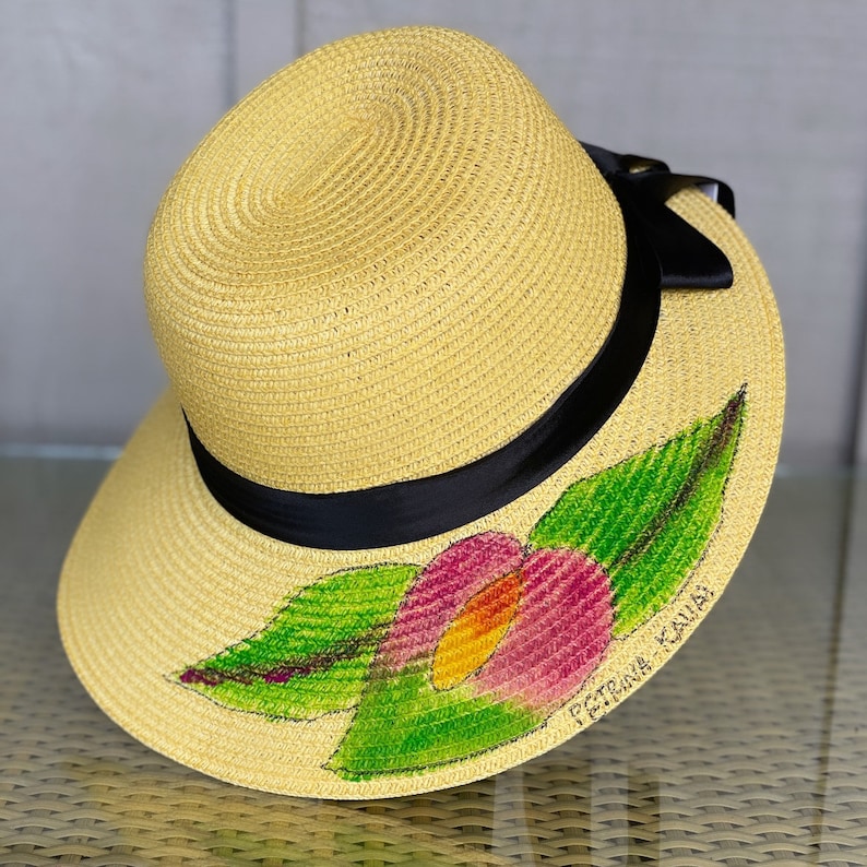 Puede incluir: Un sombrero de paja amarillo con una cinta negra alrededor de la copa. El ala presenta un dise&ntilde;o pintado a mano de una flor rosa y amarilla con hojas verdes. El texto PETRINA KAUAI est&aacute; escrito en tinta negra cerca del dise&ntilde;o floral.