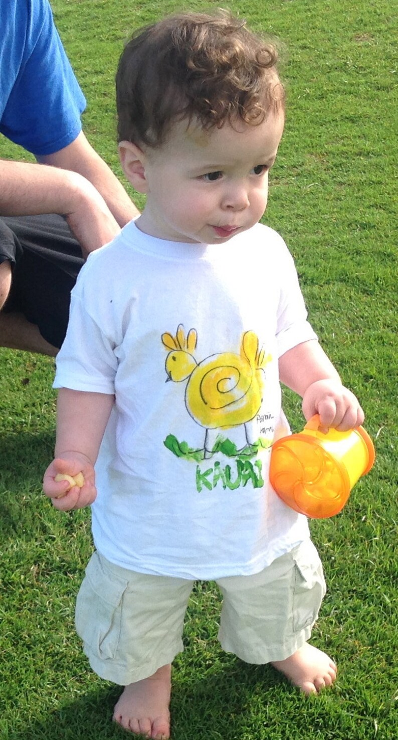 Op de afbeelding: Een jong kind draagt een wit T-shirt met een gele kipillustratie en de woorden "Pau Hana, Kauai" erop gedrukt. Het kind houdt een gele plastic beker vast.