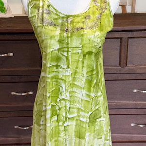 Puede incluir: Una camiseta sin mangas con un vibrante patr&oacute;n abstracto de pinceladas en verde lima y blanco. El escote est&aacute; adornado con un estampado detallado de hojas de helecho en marr&oacute;n oscuro que sigue la curva del cuello, creando un acento bot&aacute;nico.