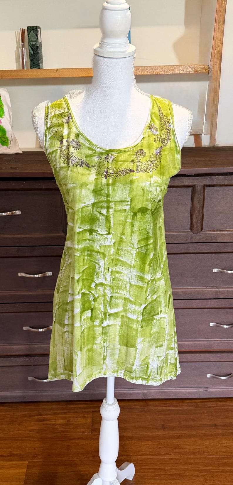 Puede incluir: Una camiseta sin mangas con un vibrante patr&oacute;n abstracto de pinceladas en verde lima y blanco. El escote est&aacute; adornado con un estampado detallado de hojas de helecho en marr&oacute;n oscuro que sigue la curva del cuello, creando un acento bot&aacute;nico.