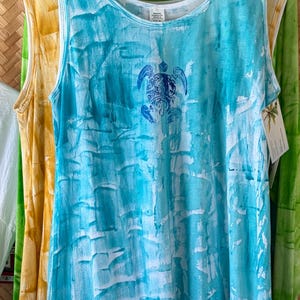 May include: A sleeveless tank top and a matching bucket hat, both featuring a white and turquoise blue tie-dye pattern. The tank top displays a blue sea turtle graphic on the chest, while the bucket hat features small blue dolphin accents.