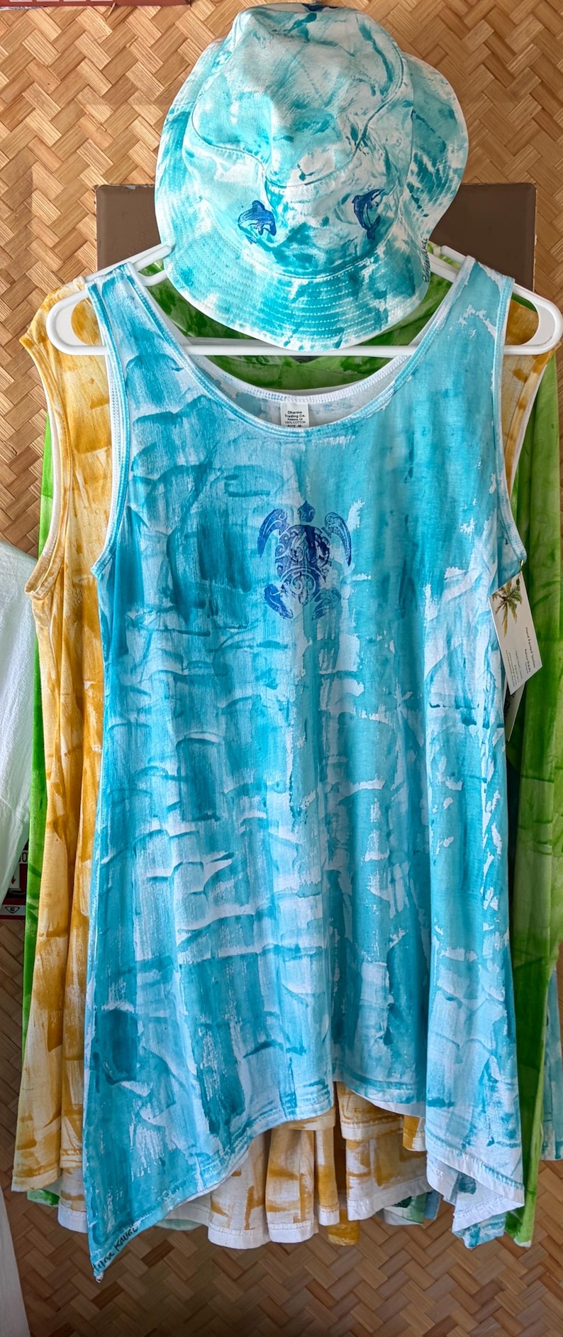 May include: A sleeveless tank top and a matching bucket hat, both featuring a white and turquoise blue tie-dye pattern. The tank top displays a blue sea turtle graphic on the chest, while the bucket hat features small blue dolphin accents.