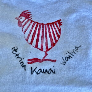 Op de afbeelding: Een wit T-shirt met een rood-wit gestreept haanontwerp. De tekst "Rehine Kauai Wailima" staat in het zwart onder de haan.