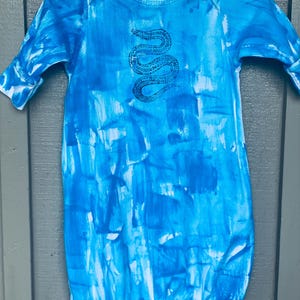 May include: A baby sleeper with a blue and white tie-dye pattern. The sleeper features a long-sleeved design and a round neckline. A black snake illustration is centered on the chest. The garment is hanging on a white hanger.