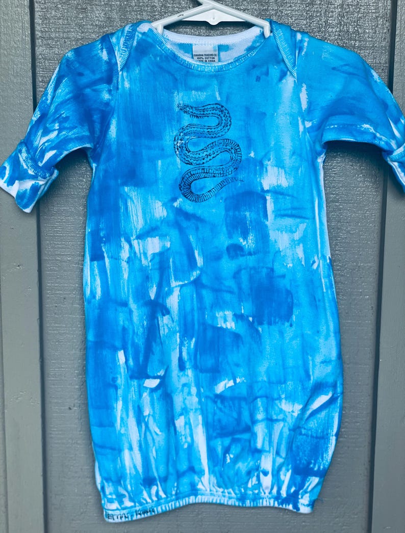 May include: A baby sleeper with a blue and white tie-dye pattern. The sleeper features a long-sleeved design and a round neckline. A black snake illustration is centered on the chest. The garment is hanging on a white hanger.