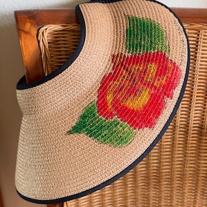 Puede incluir: Una visera de sol de paja tejida de color tostado con un borde de tela negra y un dise&ntilde;o pintado de una gran flor de hibisco roja con hojas verdes en la parte delantera. La visera cuenta con una correa de tela negra en la parte superior.