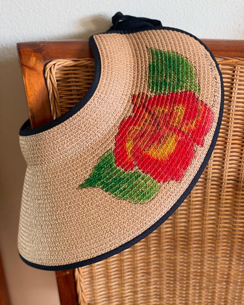 Puede incluir: Una visera de sol de paja tejida de color tostado con un borde de tela negra y un dise&ntilde;o pintado de una gran flor de hibisco roja con hojas verdes en la parte delantera. La visera cuenta con una correa de tela negra en la parte superior.