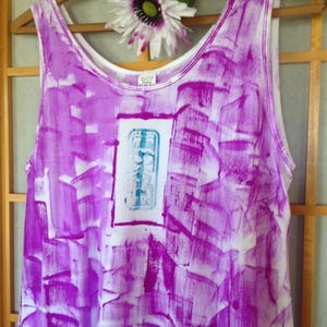 Puede incluir: Una camiseta sin mangas blanca con un vibrante patr&oacute;n de pinceladas abstractas de color p&uacute;rpura en toda la prenda. El centro muestra un panel rectangular blanco que contiene un dise&ntilde;o gr&aacute;fico azul claro con l&iacute;neas y formas verticales.