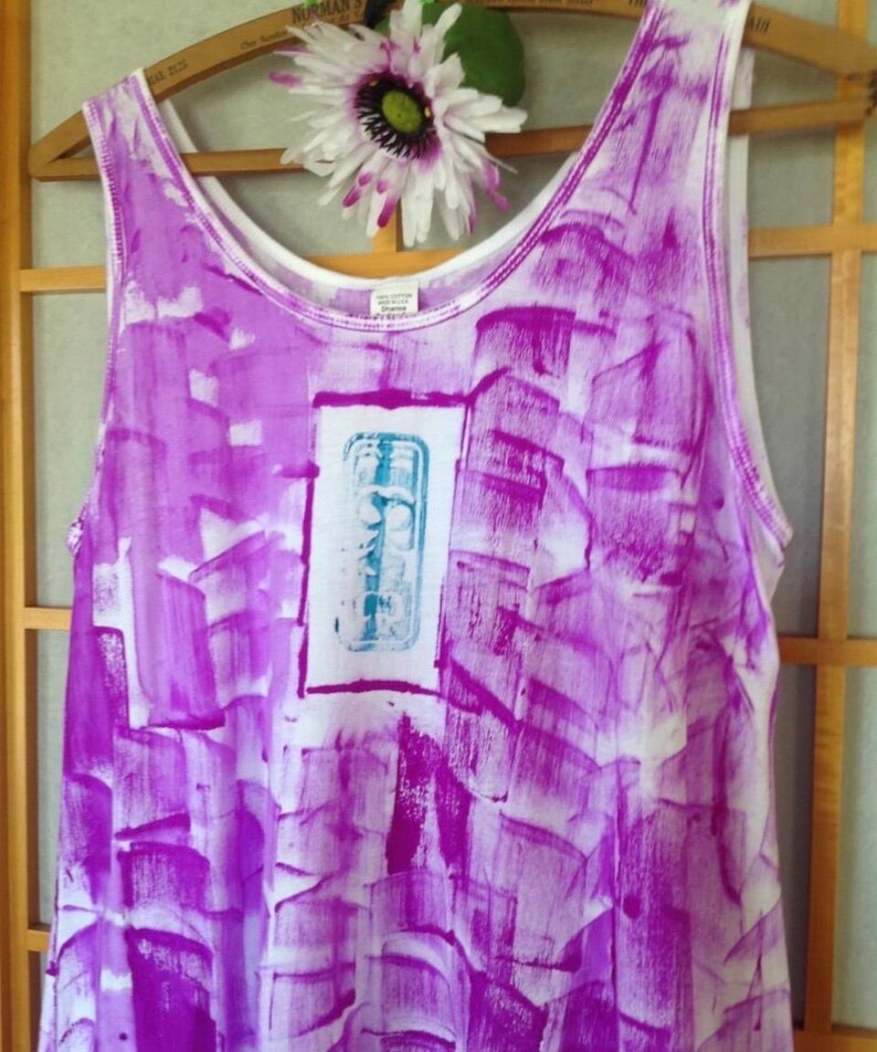 Puede incluir: Una camiseta sin mangas blanca con un vibrante patr&oacute;n de pinceladas abstractas de color p&uacute;rpura en toda la prenda. El centro muestra un panel rectangular blanco que contiene un dise&ntilde;o gr&aacute;fico azul claro con l&iacute;neas y formas verticales.