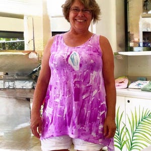 Puede incluir: Una camiseta sin mangas con un vibrante patr&oacute;n abstracto de pinceladas moradas y blancas con un acento central en forma de l&aacute;grima en blanco y azul claro, combinada con pantalones cortos blancos lisos.