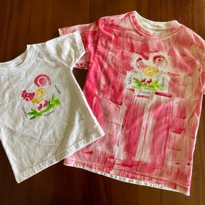 Op de afbeelding: Twee witte T-shirts met een rood haanontwerp en de woorden "Aloha", "Hawaii" en "Kauai" erop gedrukt. Een shirt is effen wit en het andere heeft een rood-wit tie-dye patroon.