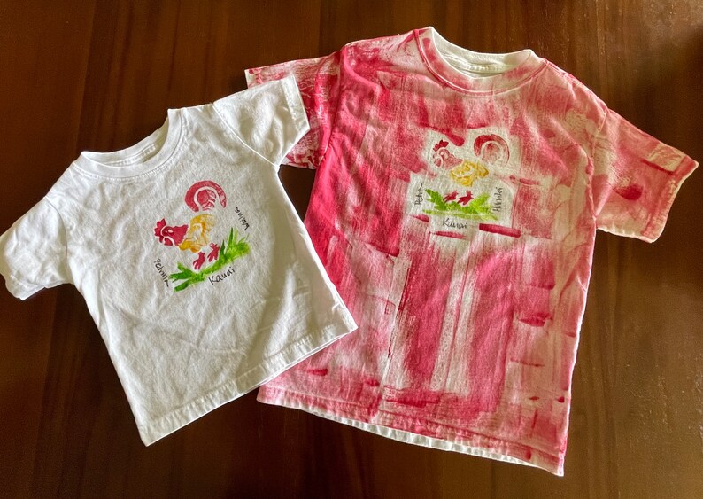 Op de afbeelding: Twee witte T-shirts met een rood haanontwerp en de woorden "Aloha", "Hawaii" en "Kauai" erop gedrukt. Een shirt is effen wit en het andere heeft een rood-wit tie-dye patroon.