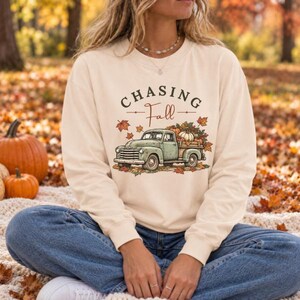 May include: A cream-colored crewneck sweatshirt featuring a graphic of a vintage light green pickup truck carrying pumpkins and autumn leaves. The text above the truck reads "CHASING Fall" in dark brown lettering, surrounded by fall foliage.