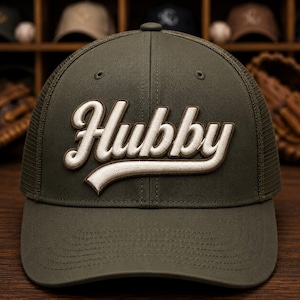 May include: An olive green baseball cap featuring a mesh back and a structured front panel. The word Hubby is embroidered in white script lettering with a dark brown outline across the centre of the front panel, featuring a curved underline.