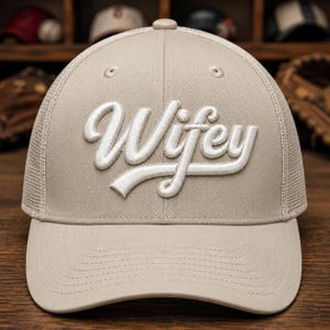 May include: A beige trucker hat featuring a mesh back and a solid front panel. The word Wifey is embroidered in white, raised script lettering across the center of the front panel. The hat has a curved brim and a button at the top center.