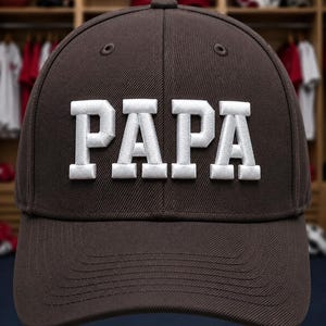 May include: A dark brown baseball cap featuring the word PAPA embroidered in large, bold, white block letters across the front panels. The cap has a structured crown, a curved brim, and stitched eyelets on the top panels for ventilation.