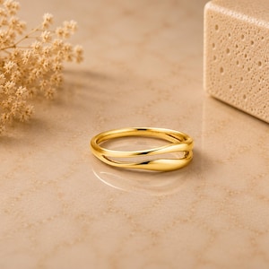 May include: A polished gold ring featuring a smooth, open-work design with two parallel bands that curve and meet at the sides. The ring has a high-shine metallic finish and a continuous, fluid shape that creates a small gap between the two bands.