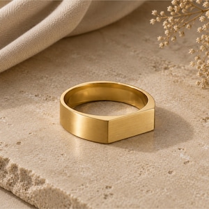 May include: A gold-toned metal ring featuring a flat, rectangular top surface and a smooth, polished band. The ring has a solid construction with a consistent metallic finish, showcasing a clean, geometric design with a prominent, flat front face.