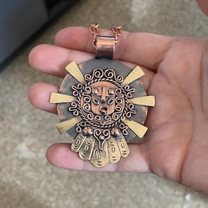 May include: A copper-colored pendant with a detailed, sun-like design. The pendant features a central face with intricate patterns, surrounded by radiating gold-colored elements. The pendant is suspended from a copper-colored chain.