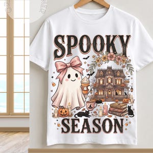 May include: A white short-sleeve t-shirt featuring a graphic print with the text SPOOKY SEASON. The design includes a ghost with a pink bow holding a coffee cup, a haunted house, pumpkins, candles, books, and black cats in a muted color palette.