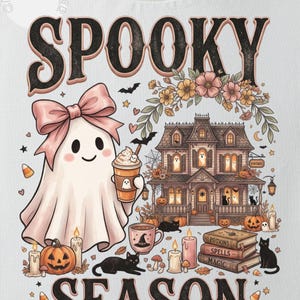 May include: A white t-shirt featuring a graphic print with the text SPOOKY SEASON. The design includes a ghost wearing a pink bow holding a coffee cup, a large house, black cats, pumpkins, candles, and a stack of books labeled POTIONS, SPELLS, and MAGIC.