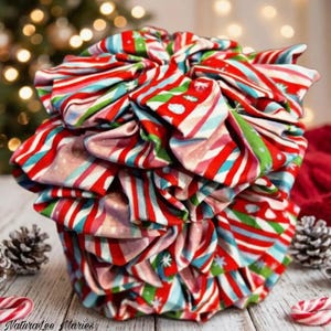 May include: A large, ruffled fabric hair scrunchie featuring a repeating pattern of diagonal red, white, green, and light blue stripes with small white snowflake accents. The scrunchie has a voluminous, gathered texture.