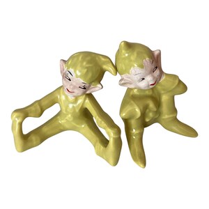May include: Two ceramic elf figurines with a glossy yellow finish. Each elf features pointed ears, a matching yellow hat, and painted facial details including red lips and dark eyelashes. The figures are posed in seated positions.