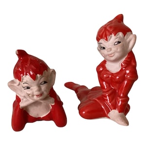 May include: Two ceramic elf figurines with glossy red clothing and pointed hats. One elf is lying down on its stomach, while the other is sitting upright with its legs extended. Both figures have pale skin, pointed ears, and painted facial features.