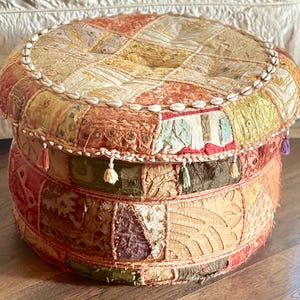 May include: A round, low-profile ottoman featuring a patchwork design of various fabrics in shades of orange, yellow, green, and brown. The top and sides are decorated with small white cowrie shells and small, colorful fabric tassels.