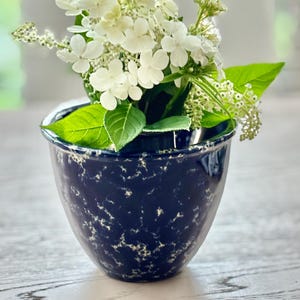 Può includere: Un vaso da fiori in ceramica blu navy profondo con un motivo bianco maculato contenente un bouquet di ortensie bianche e foglie verdi. Il vaso ha una forma arrotondata e affusolata con una finitura liscia e lucida e un bordo superiore leggermente svasato.