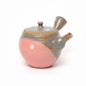 Peut inclure: Une théière kyusu en céramique avec un corps arrondi et une finition bicolore. La partie inférieure est rose mat uni, tandis que la partie supérieure, le couvercle et la poignée latérale présentent un vernis gris et brun texturé et tacheté.