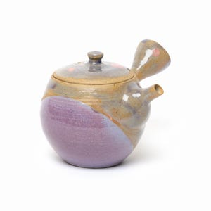 May include: A ceramic teapot with a side handle and a lid, featuring a rounded body with a two-tone glaze. The lower portion is a solid lavender purple, while the upper section and lid are a mottled tan and grey with small pink accents.
