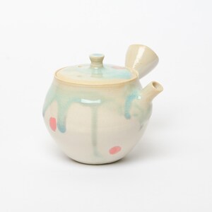 Peut inclure: Une théière kyusu en céramique avec une anse latérale et un couvercle. Le corps est blanc cassé avec une glaçure bleu clair qui coule depuis le bord supérieur. De petites taches roses circulaires sont dispersées sur la surface.