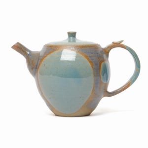 Peut inclure: Une théière en céramique présentant un corps arrondi avec un centre circulaire émaillé bleu clair, contrasté par une teinte de base terreuse et beige chaud. Elle comprend un couvercle assorti avec un petit bouton, un bec verseur et une anse.