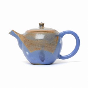 May include: A ceramic teapot featuring a rounded body with a glossy blue glaze on the lower section and a textured, earthy brown finish on the upper portion and lid. The teapot includes a curved handle and a short, angled spout for pouring.