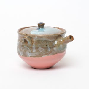 Peut inclure: Une théière en céramique avec un corps arrondi, une base rose mate et une partie supérieure en terre cuite texturée ornée de coups de pinceau blancs. Elle comprend un bec verseur latéral, une petite anse et un couvercle assorti émaillé bleu.