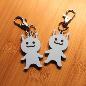 May include: Two light blue keychains shaped like small alien figures with two antennae on their heads. Each figure features a simple dark brown printed face with two round eyes and a curved mouth. A silver metal lobster clasp is attached to each.