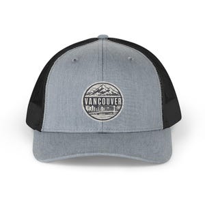 May include: A heather gray trucker hat with a black mesh back and a circular patch on the front. The patch features a black and white graphic of mountains, a city skyline, and a bridge, with the word VANCOUVER printed in the center of the design.