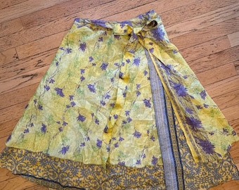 Può includere: Una gonna a portafoglio con base gialla e stampa floreale viola. La gonna presenta una chiusura a fiocco in vita e un orlo a strati che rivela un tessuto a contrasto con motivo grigio e giallo sotto lo strato floreale principale.