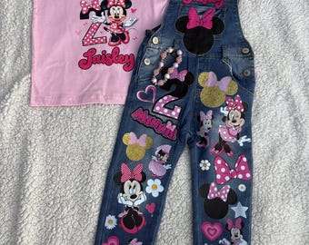 Może przedstawiać: Jasnoróżowy t-shirt z napisem Oh, Toodles i Saisley oraz grafiką Myszki Minnie, w zestawie z niebieskimi ogrodniczkami dżinsowymi z naszywkami Myszki Minnie, imieniem Saisley, cyfrą 2 oraz różowym wykończeniem ze sztucznego futra przy ramiączkach i nogawkach.