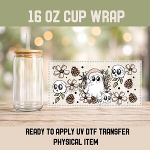 May include: A clear glass tumbler with a bamboo lid and straw next to a rectangular UV DTF transfer sheet. The sheet features a pattern of white ghosts wearing Santa hats, skulls, brown pinecones, and light pink flowers on a cream background.