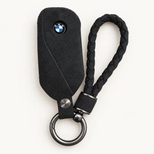 Puede incluir: Una funda de ante negro para llave de coche BMW con el emblema circular de BMW, un remache metálico plateado y una correa de muñeca trenzada de ante negro a juego, unida a un llavero circular de metal oscuro.