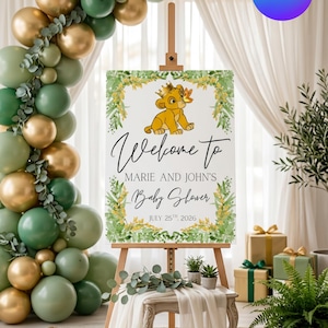May include: A rectangular baby shower welcome sign on a wooden easel featuring a cartoon lion cub wearing a crown, green foliage, and yellow flowers. The text reads: Welcome to MARIE AND JOHN'S Baby Shower JULY 25TH, 2026. The sign is framed by balloons.