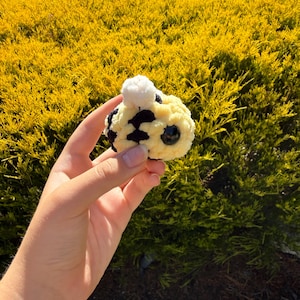 May include: A small, round, crocheted bee plush toy featuring a soft yellow and black striped body, a white pom-pom accent on top, and a single large, smooth, black plastic eye visible on the side of its textured, yarn-based spherical form.