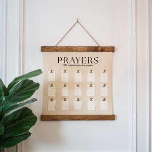 May include: A wall-hanging prayer board featuring a cream-colored canvas with the text PRAYERS and little prayers become big miracles in black font. It includes fifteen small white paper cards attached with gold clips and dark wood rails.