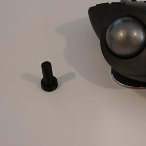 May include: A black ergonomic trackball mouse featuring a large metallic gray ball positioned on the side, multiple buttons along the top, and a silver base. Beside the mouse sits a small, black, cylindrical plastic component with a flat top.