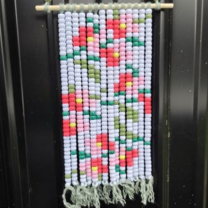 May include: A rectangular wall hanging made of white, pink, red, yellow, and green plastic beads arranged in a floral pattern. The beads are strung on green yarn attached to a horizontal wooden dowel with green beads and a hanging loop.